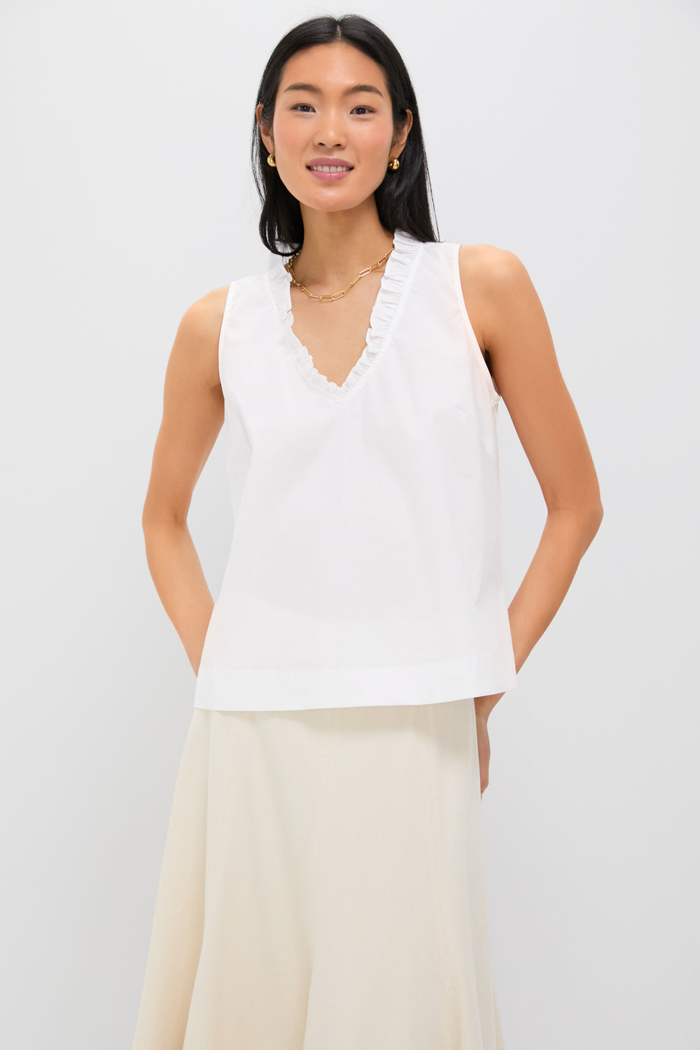 A woman wears the Pomander Place Veronica Top, a sleeveless ruffle blouse with a V-neckline and subtle trim, paired with a light-colored skirt.