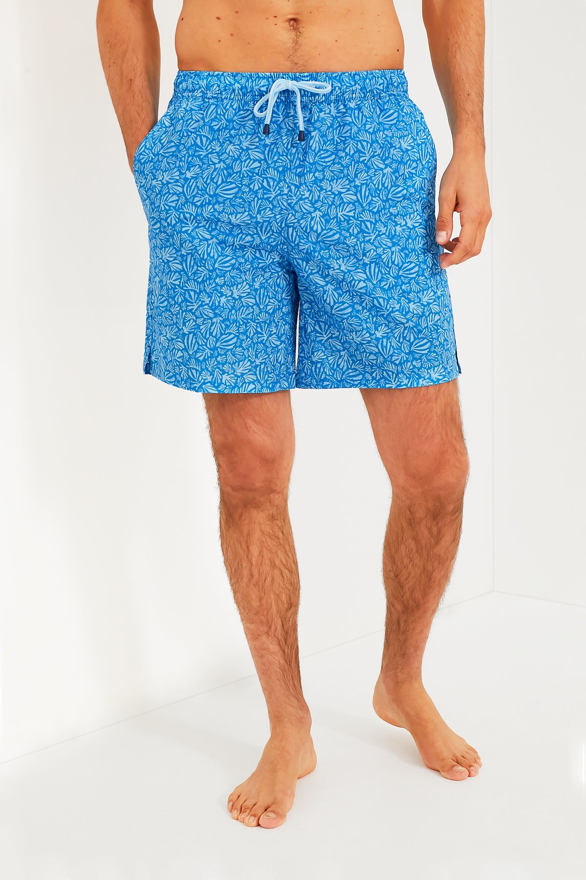 Man stands barefoot with hands in pockets, wearing blue patterned Peter Millar swim trunks. These quick-drying trunks offer four-way stretch for extra comfort.