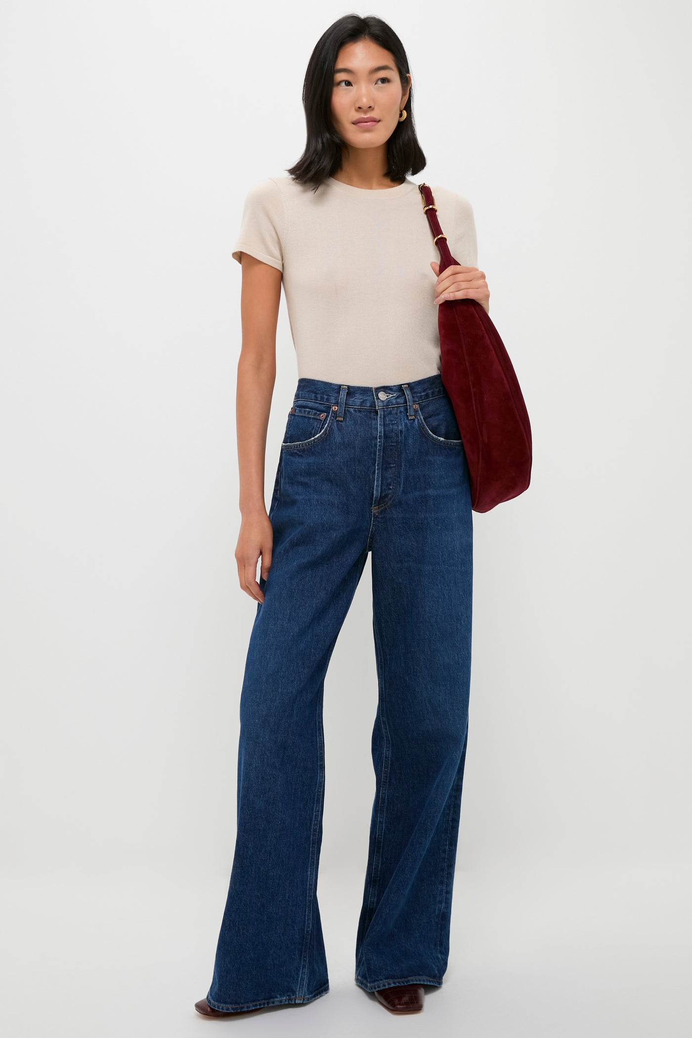 A woman poses against a plain backdrop wearing a beige short-sleeve top, high-waisted wide-leg blue jeans, brown shoes, and carries a red Liffner crescent hobo bag.