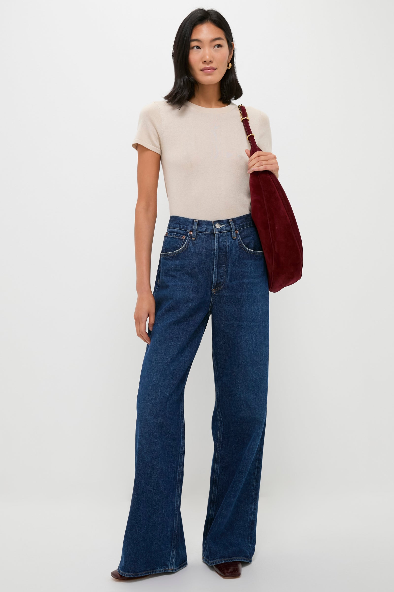 A woman poses against a plain backdrop wearing a beige short-sleeve top, high-waisted wide-leg blue jeans, brown shoes, and carries a red Liffner crescent hobo bag.