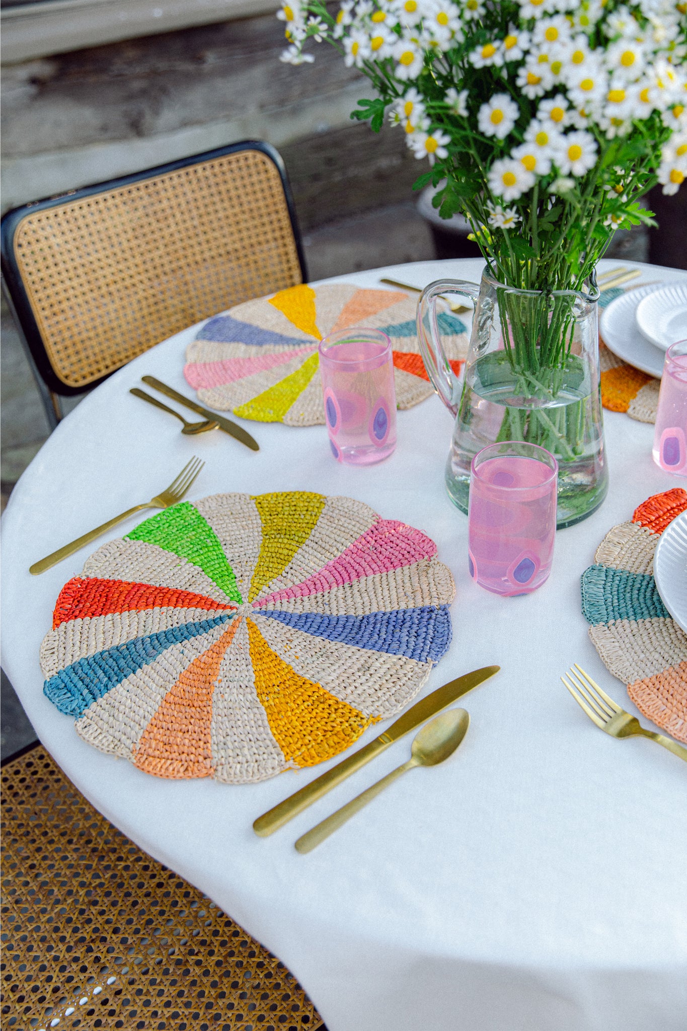 Payton James round table set with handmade colorful decor, raffia placemats, gold cutlery, pink glasses, white plates, and a daisy-filled pitcher on a white tablecloth.