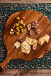Teak Puro Round Serving Board elegantly displays a classic, effortless snack mix of olives, nuts, figs, grapes, and flatbread, embodying high-end, fun entertaining.