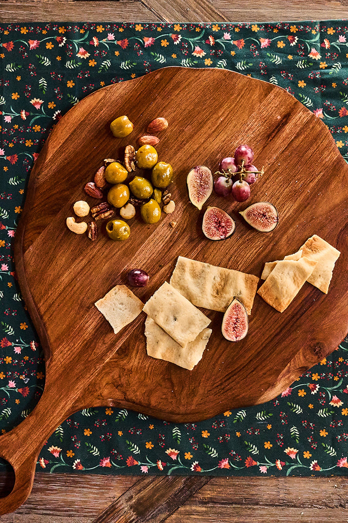 Teak Puro Round Serving Board elegantly displays a classic, effortless snack mix of olives, nuts, figs, grapes, and flatbread, embodying high-end, fun entertaining.