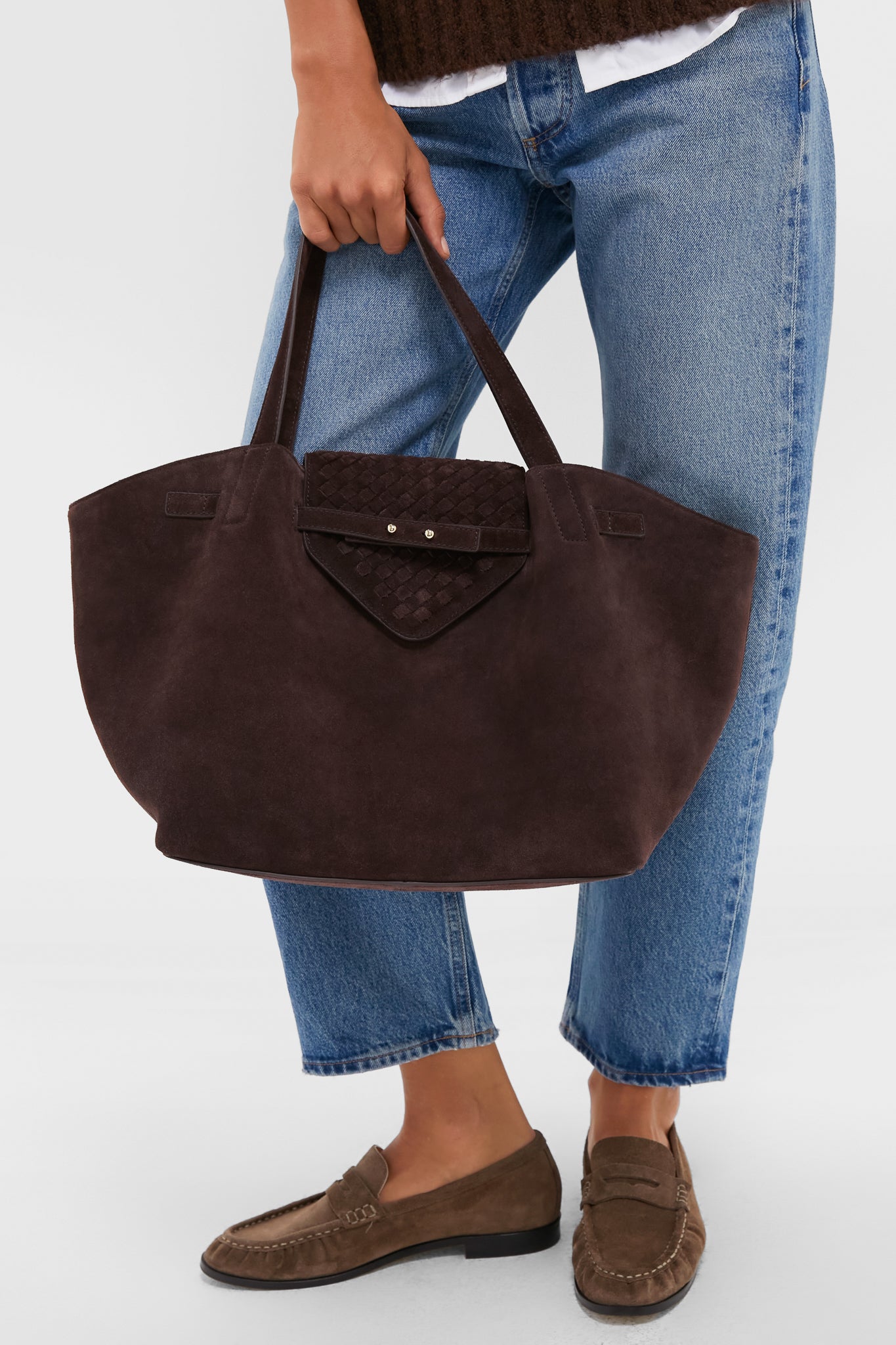 A person in blue jeans and brown loafers carries a large, dark brown suede work tote by Tuckernuck Accessories, featuring two handles and a front flap pocket—ideal for daily essentials or use as a stylish laptop bag.