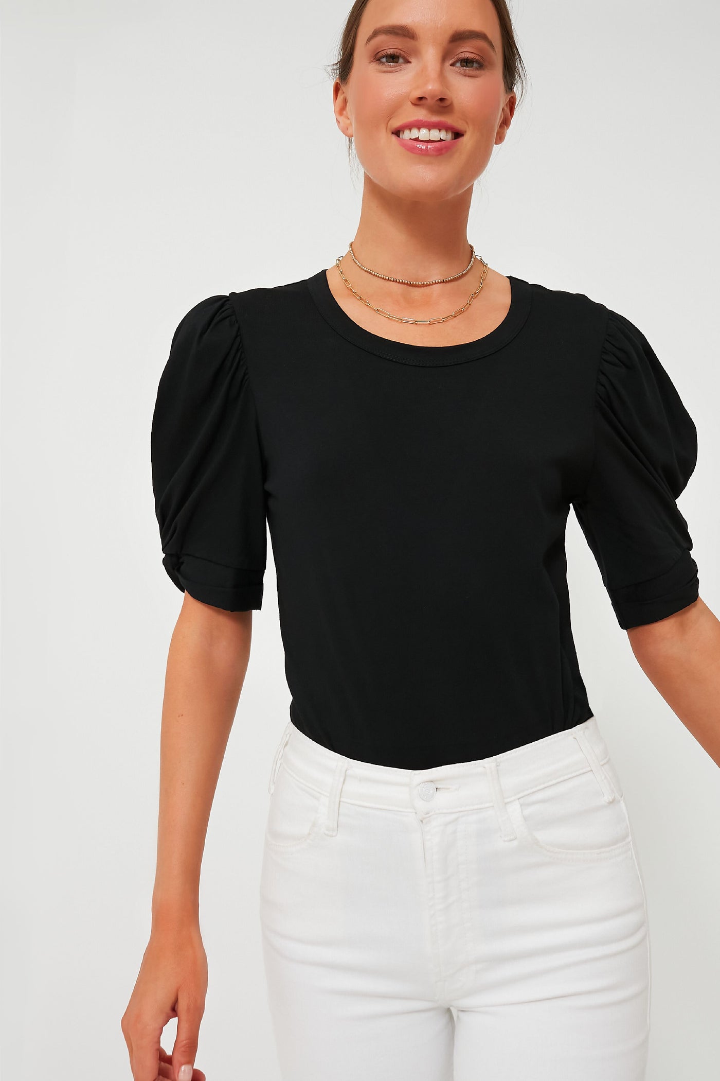 A woman in a Pomander Place Black Vesty Puff Sleeve Crewneck, layered gold necklaces, and high-waisted white jeans stands confidently, smiling.