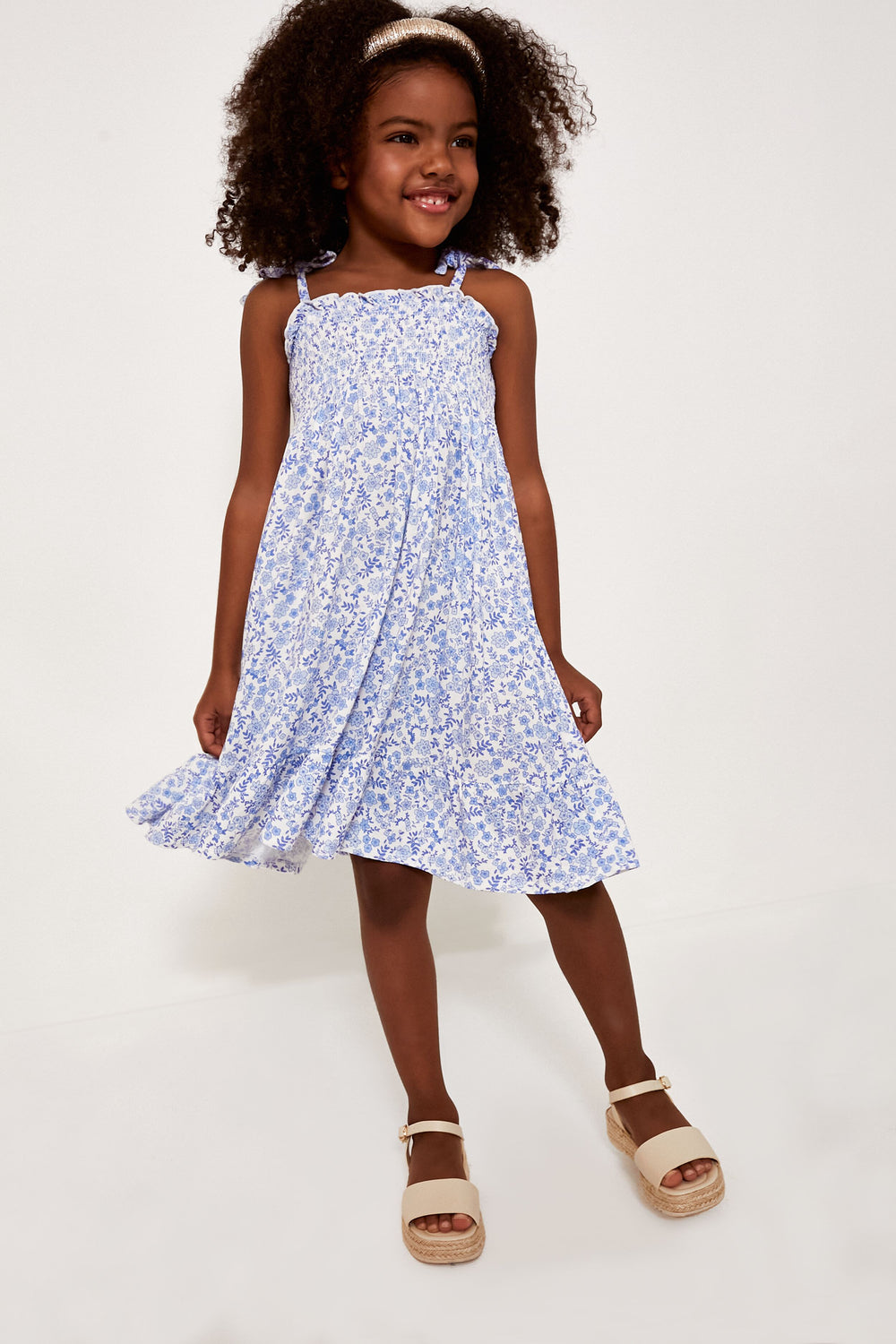 A young girl wearing an Angel Dear light blue floral dress and beige sandals, smiling and looking slightly to the side.