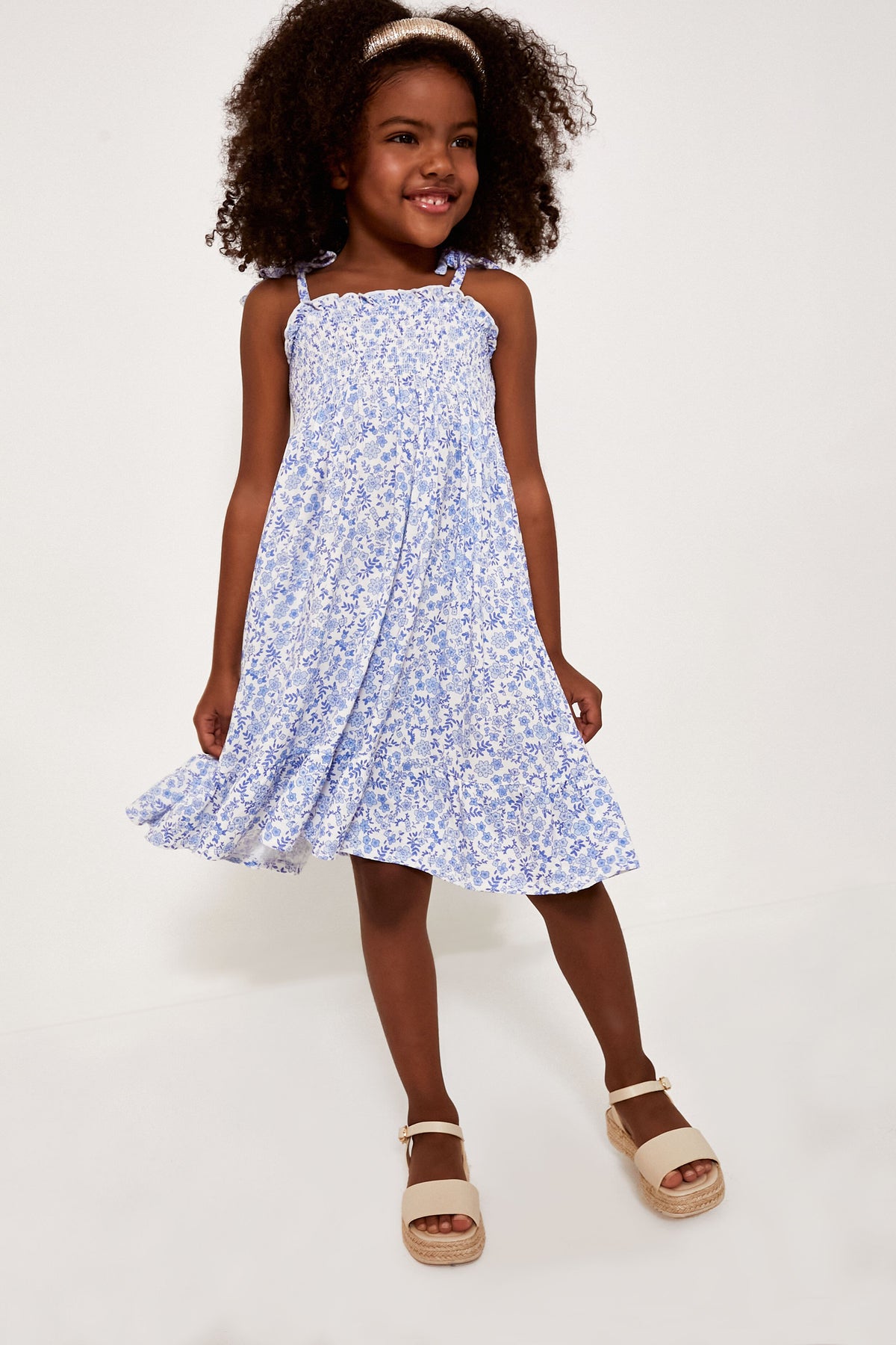 A young girl wearing an Angel Dear light blue floral dress and beige sandals, smiling and looking slightly to the side.