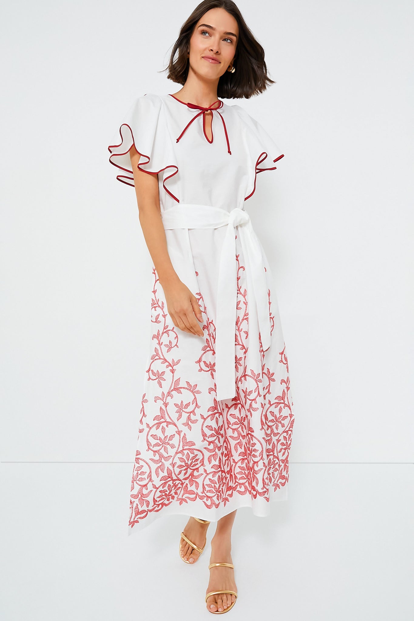 A woman wears the Loretta Caponi Strawberry Leaves Daisy Dress, an embroidered maxi with red floral details, flutter sleeves, waist tie, and flat sandals for a feminine touch.