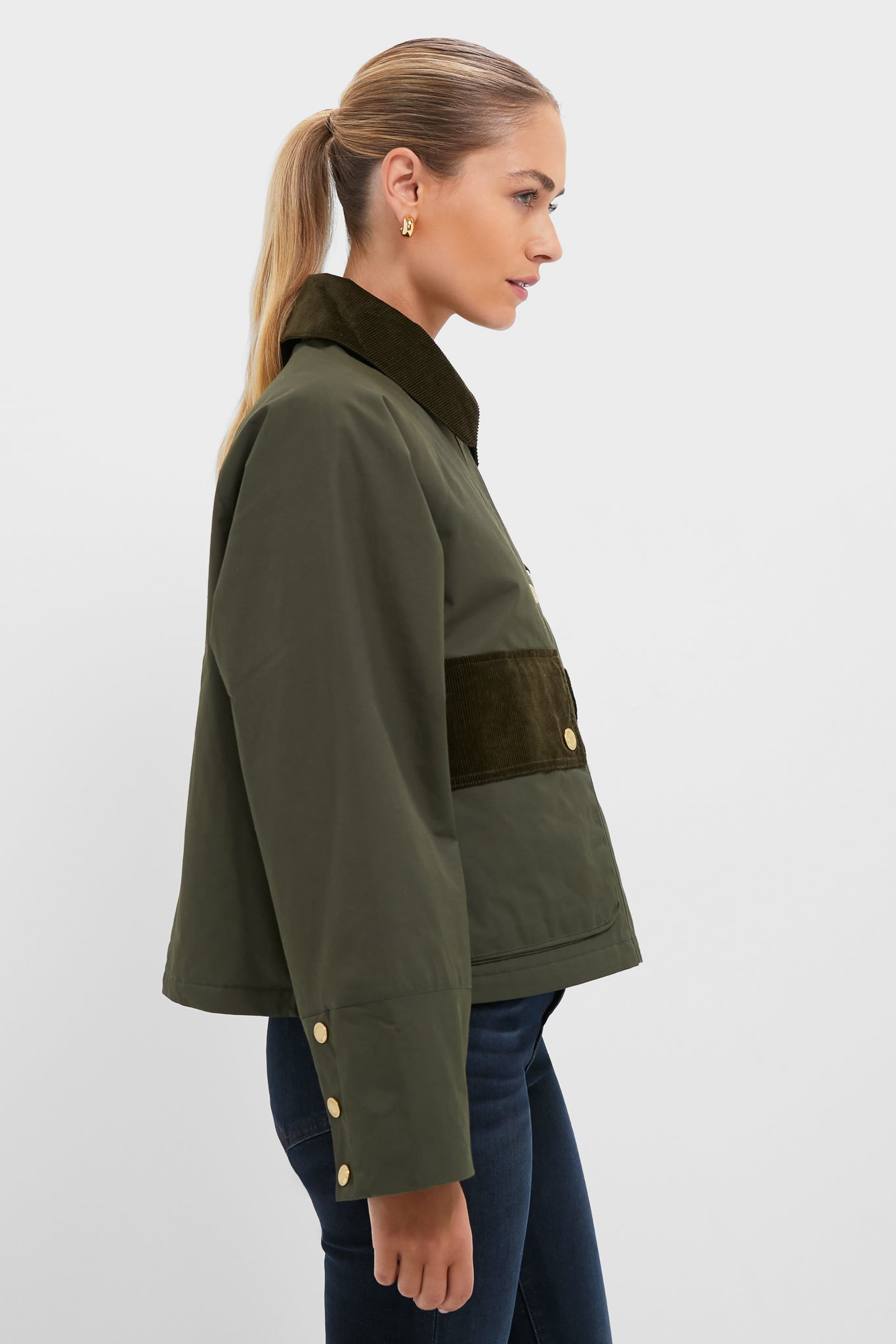 A woman in side profile wears the Barbour Olive Phoebe Casual Jacket with a dark green corduroy collar and accents, paired with dark blue jeans, against a plain background.