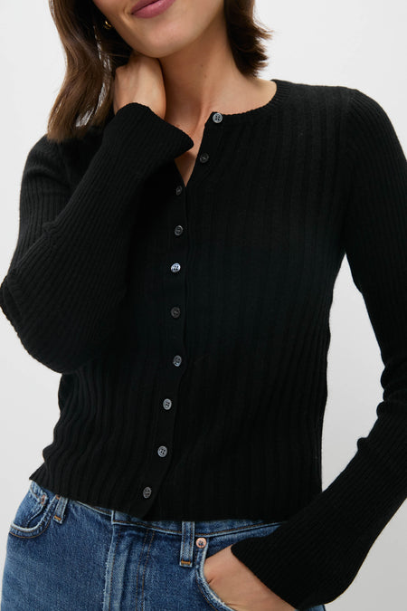 A woman wearing an Alex Mill black ribbed cotton-wool blend fitted cardigan and blue jeans stands, touching her neck with one hand.
