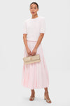 A woman wears a textured knit white short-sleeve top by molli, a pale pink pleated midi skirt, nude heeled sandals, and carries a woven beige clutch.