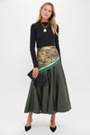 A woman in a Tuckernuck black ultra-soft knit long-sleeve top with high crewneck, green A-line skirt, floral waist scarf, black heels, and a clutch poses against a plain white background.