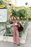 Woman waving in front of a metro station in a pink striped blouse and matching pants. 