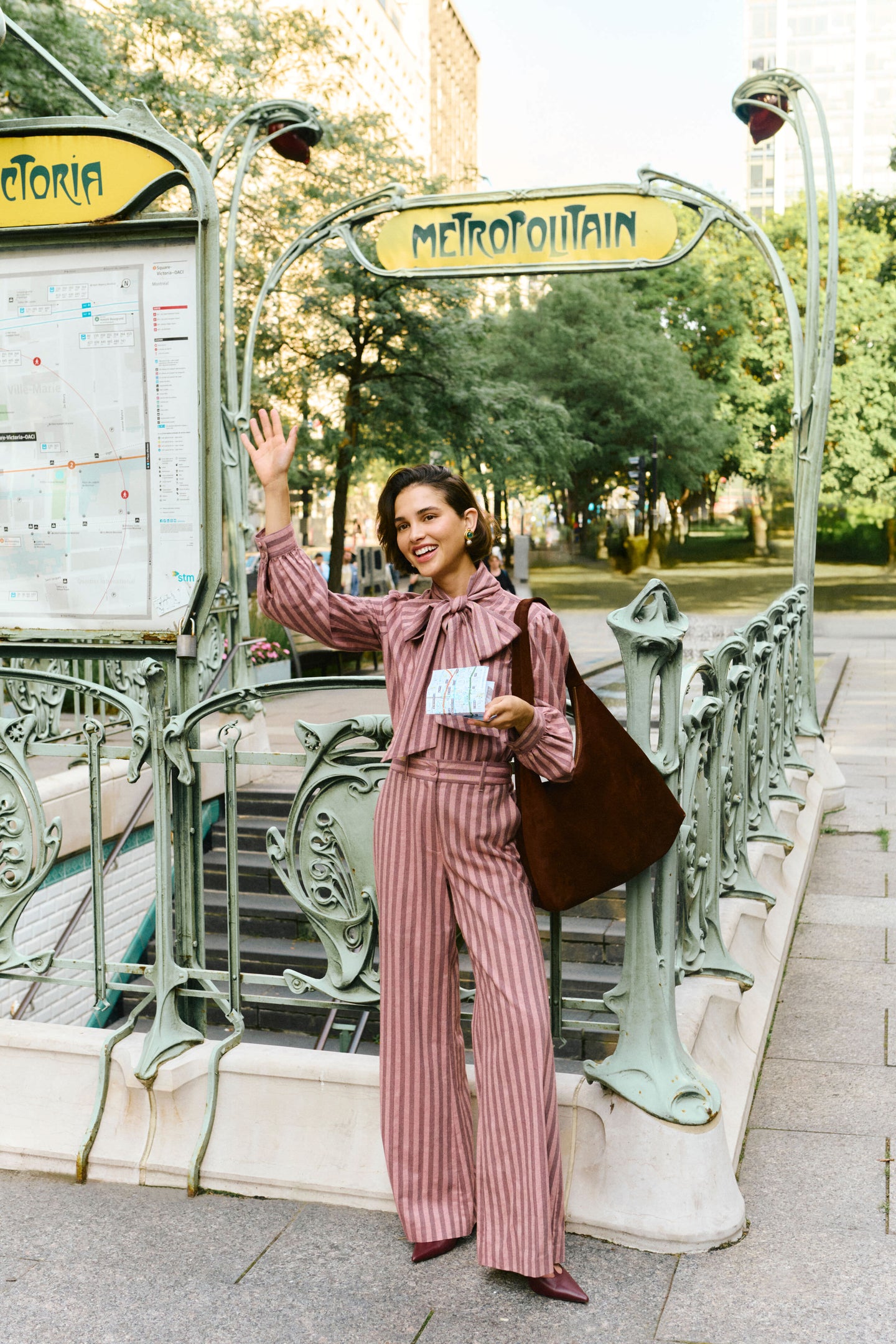 Woman waving in front of a metro station in a pink striped blouse and matching pants. 