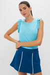A woman wears a Tnuck Sport light blue athleisure tank with ruffled shoulders and quick-dry fabric, paired with a dark blue skirt featuring white vertical stripes.