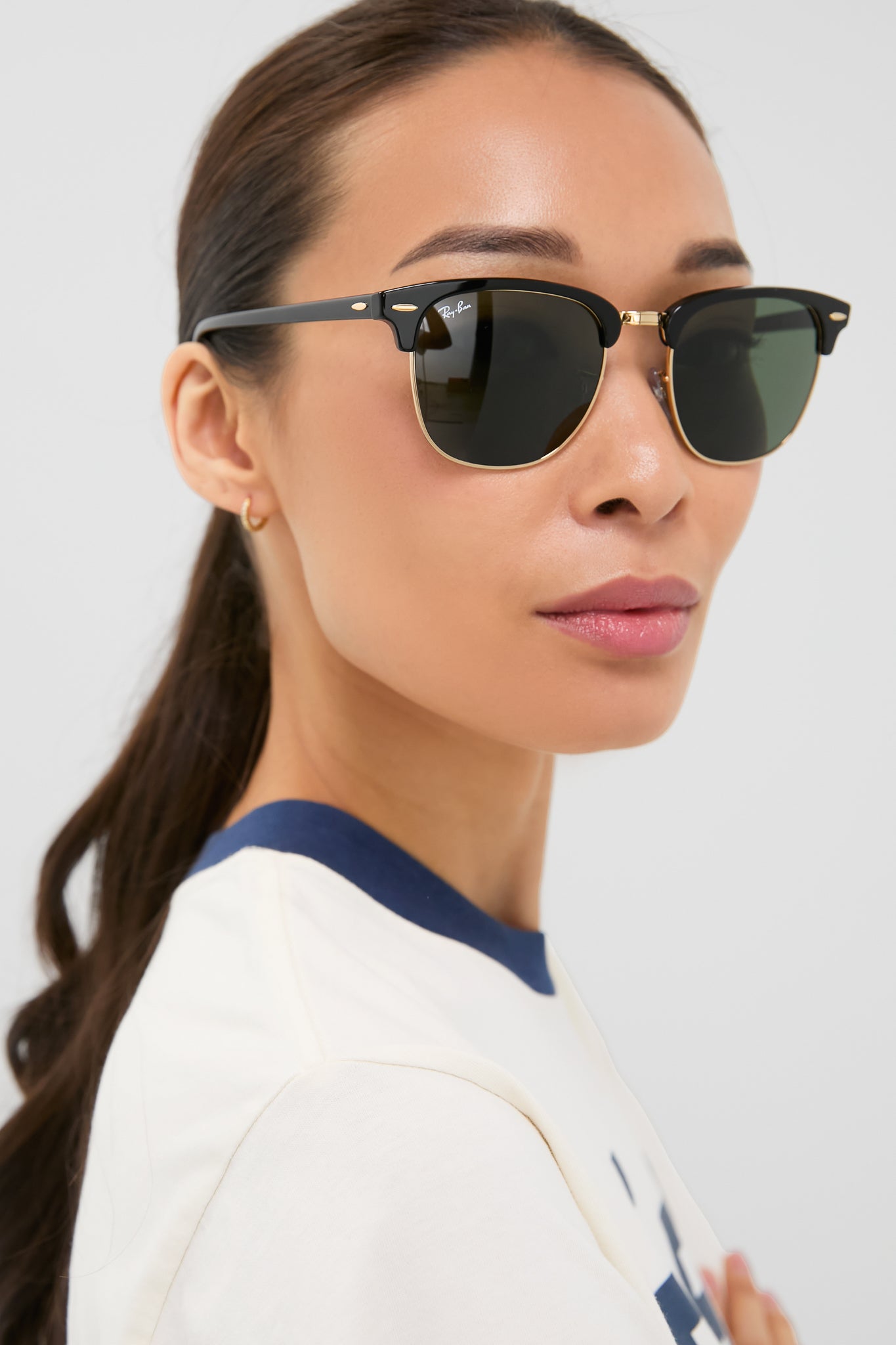 A woman with long dark hair in a ponytail wears Ray-Ban sunglasses and a cream-colored top with blue trim, glancing over her shoulder against a plain backdrop.