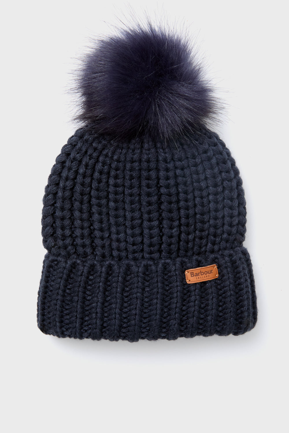 A chunky-knit navy blue Barbour beanie featuring a large faux-fur pom-pom on top and a small brown Barbour logo patch on the front cuff.