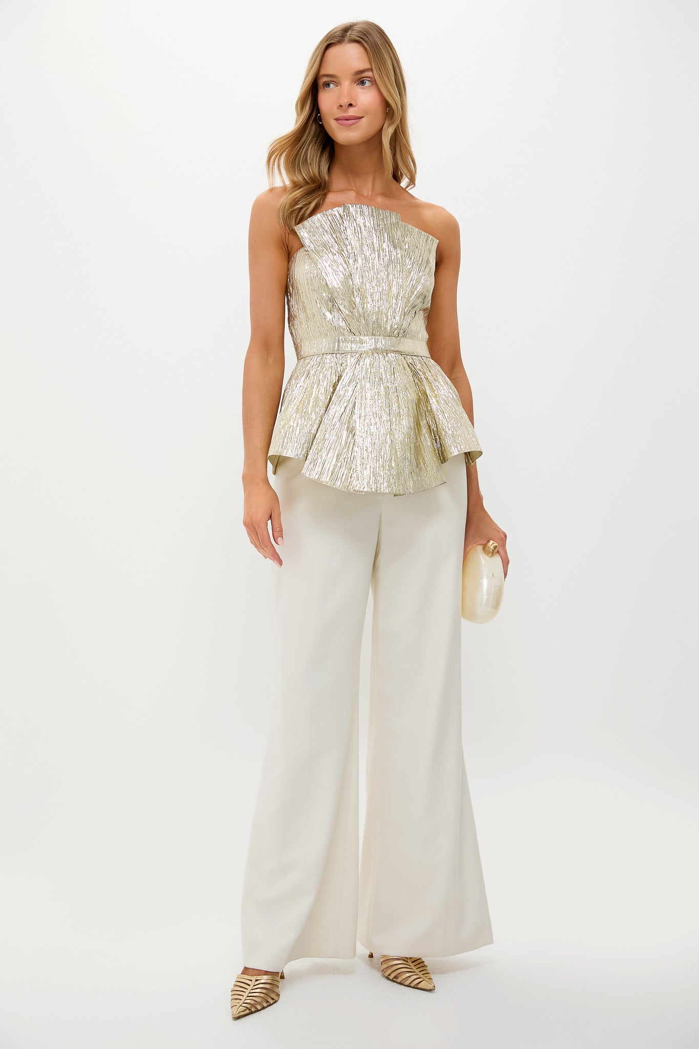 Model wearing the Gold Plisse Iris Strapless Top, a classic, high-end metallic gold piece styled effortlessly with ivory trousers, clutch, and strappy mules for a fun, elegant look.