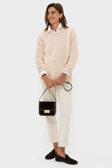 A woman wears a beige crewneck sweater over a white collared shirt, cream pants, and black loafers, styled with a small black handbag.