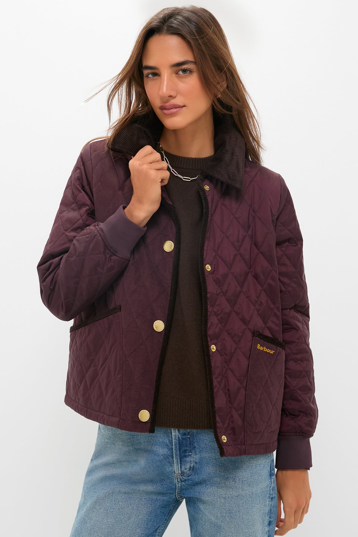 A woman wearing a black cherry Barbour Marla Quilted Jacket with gold buttons over a brown sweater and blue jeans stands against a plain white background.