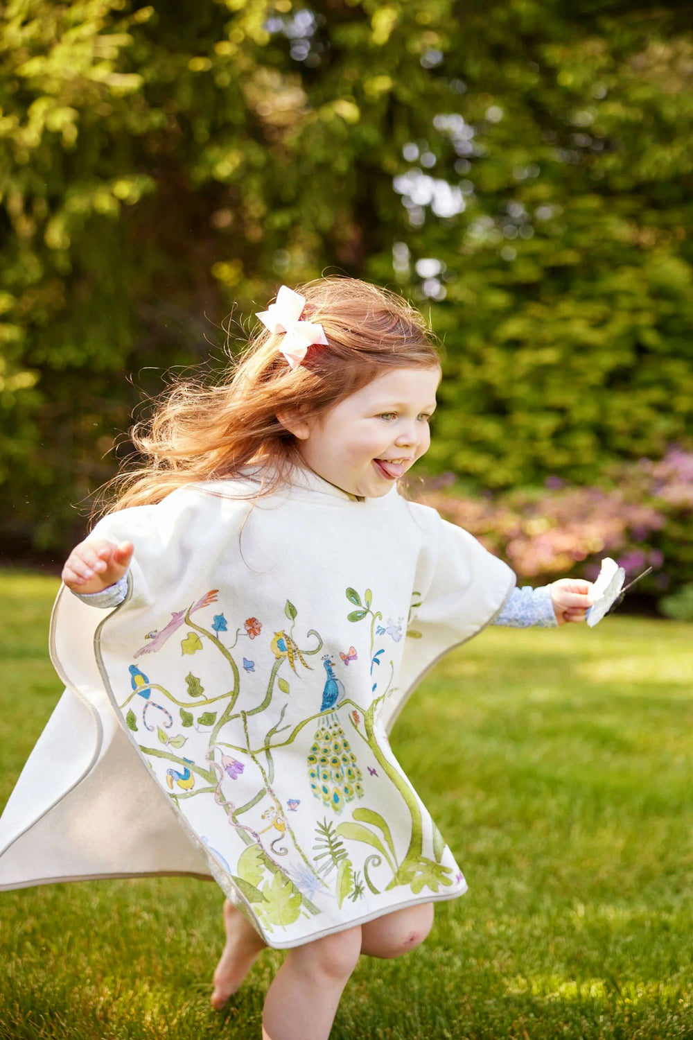 A young girl runs barefoot on green grass, wrapped in the comfort of a Timo & Violet heirloom-quality white organic cotton terry cape featuring colorful bird and plant designs.