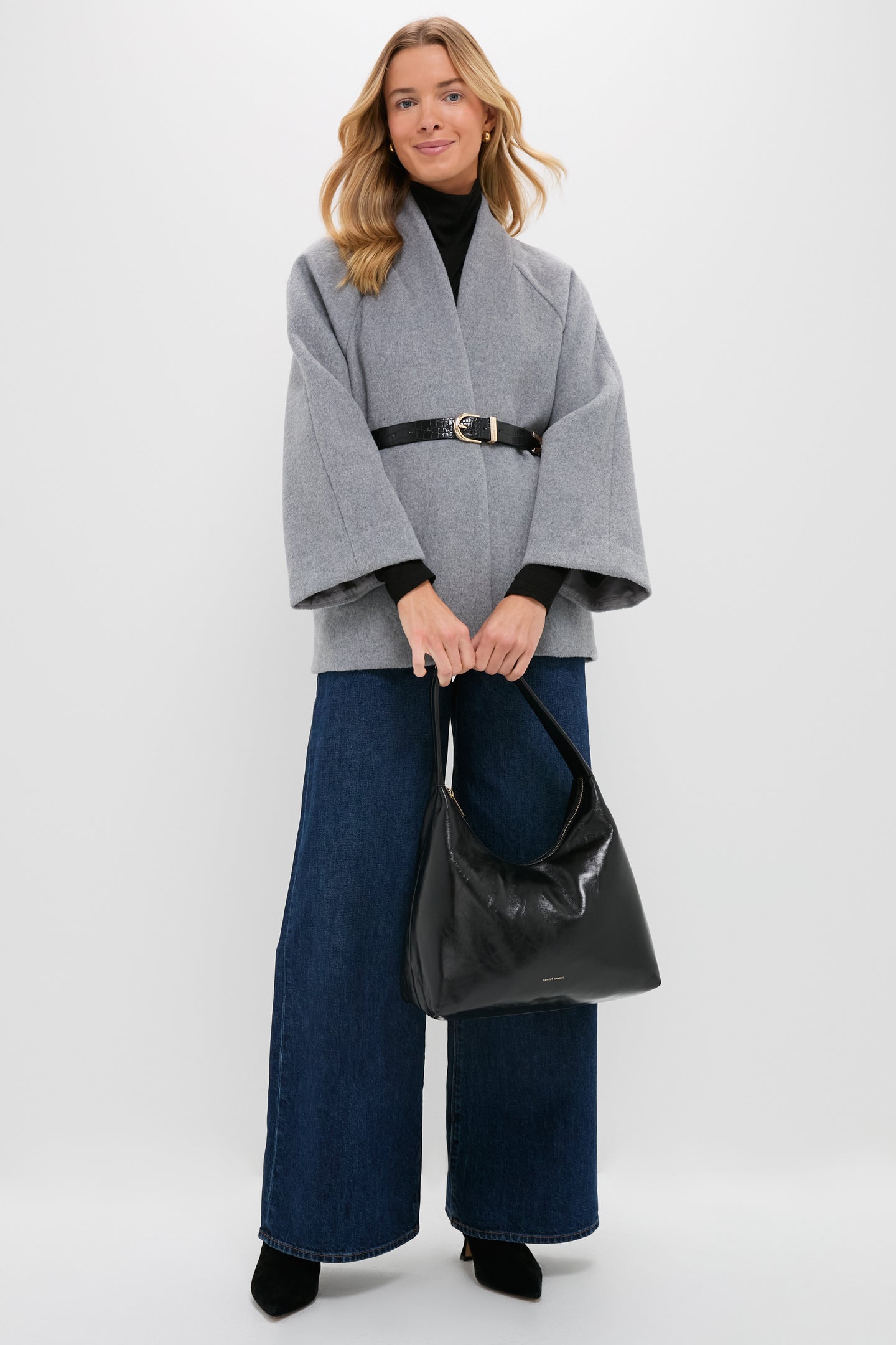 A woman stands against a plain background wearing a gray belted coat, wide-leg blue jeans, black shoes, and holding a Mansur Gavriel Candy Hobo laminated leather handbag.