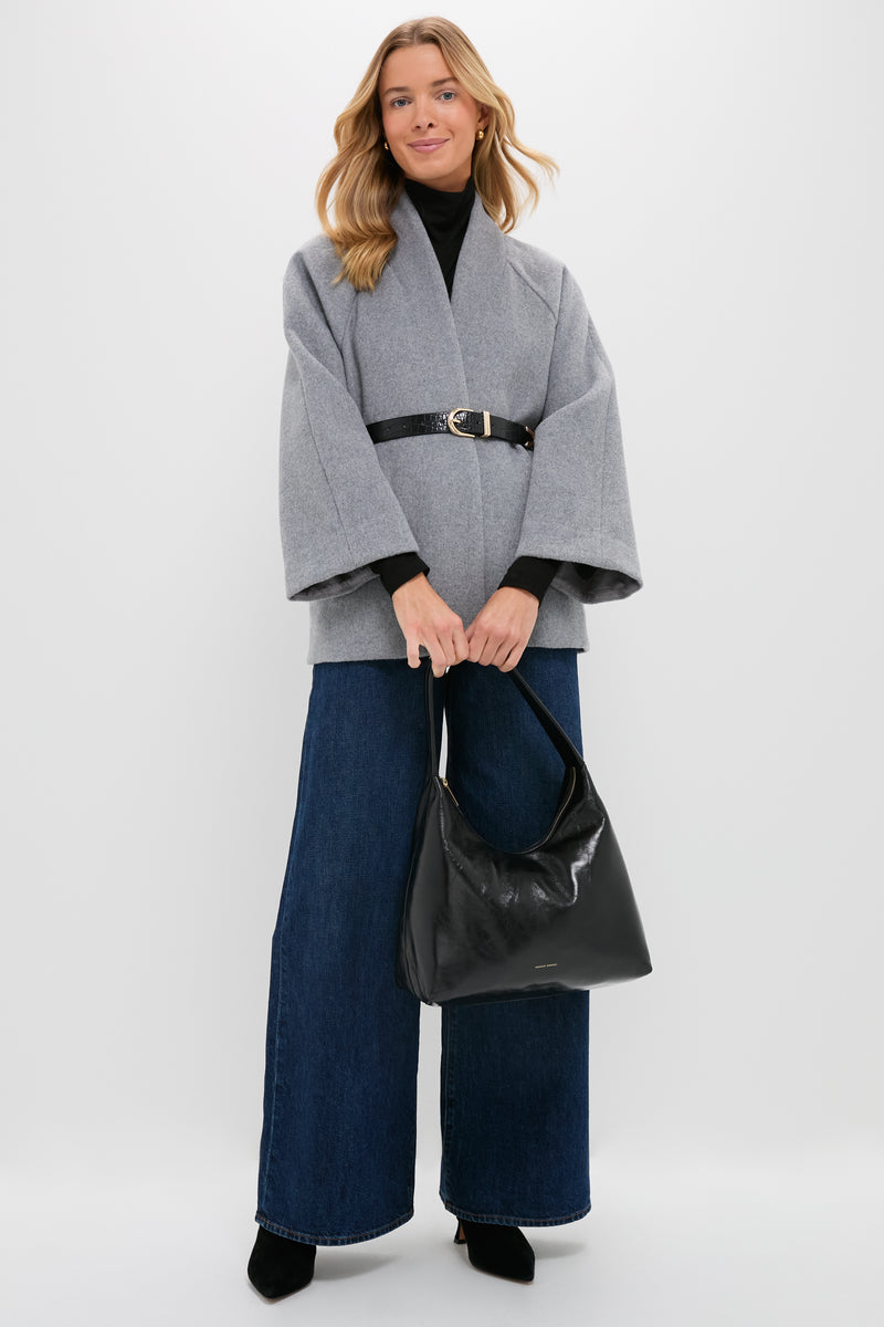 A woman stands against a plain background wearing a gray belted coat, wide-leg blue jeans, black shoes, and holding a Mansur Gavriel Candy Hobo laminated leather handbag.