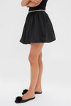 Black Taffeta Embellished Camila Mini Skirt, featuring a bubble hem and rhinestone belt, paired effortlessly with black pom-pom flats for a classic, high-end look.