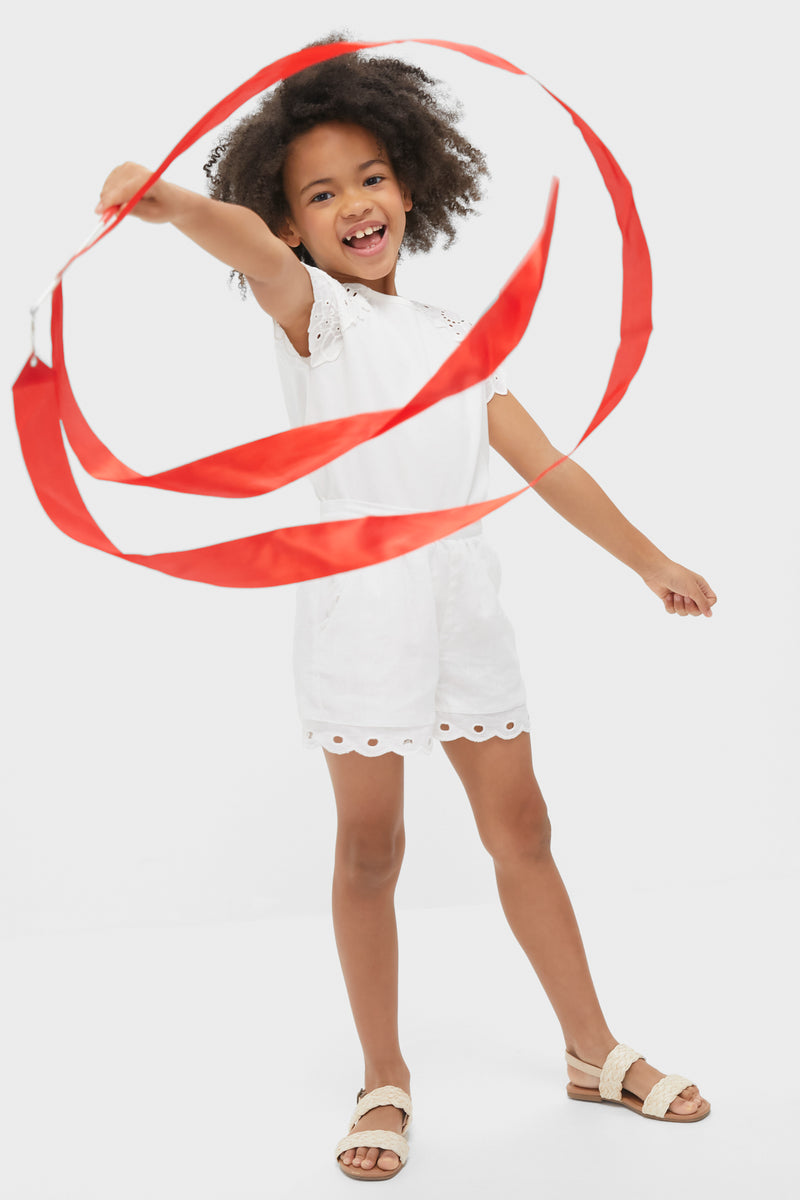 A young girl wears a white eyelet sleeve blouse, styled with white shorts and white sandals while waving a long red ribbon.
