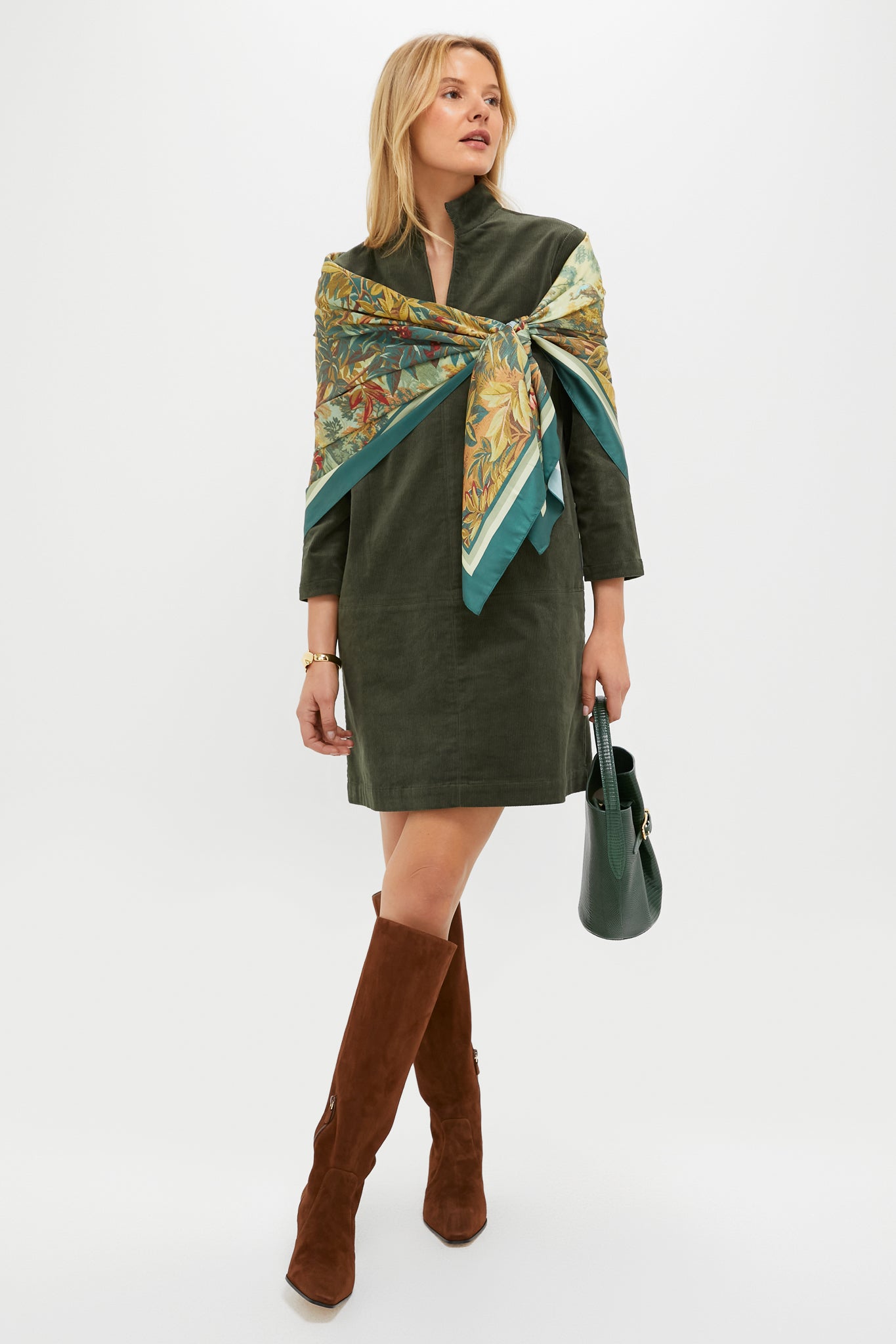 A woman stands against a plain white background, wearing a dark green dress, patterned scarf, brown knee-high Stuart Weitzman boots, and holding a green handbag.