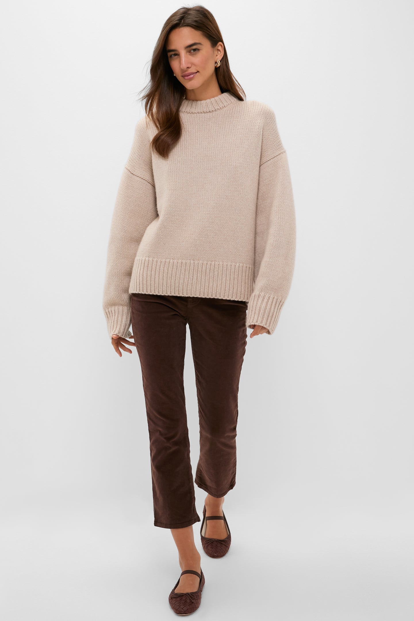 A woman stands against a plain light background wearing a Tuckernuck beige cashmere boxy-fit sweater, brown cropped pants, and brown flats.