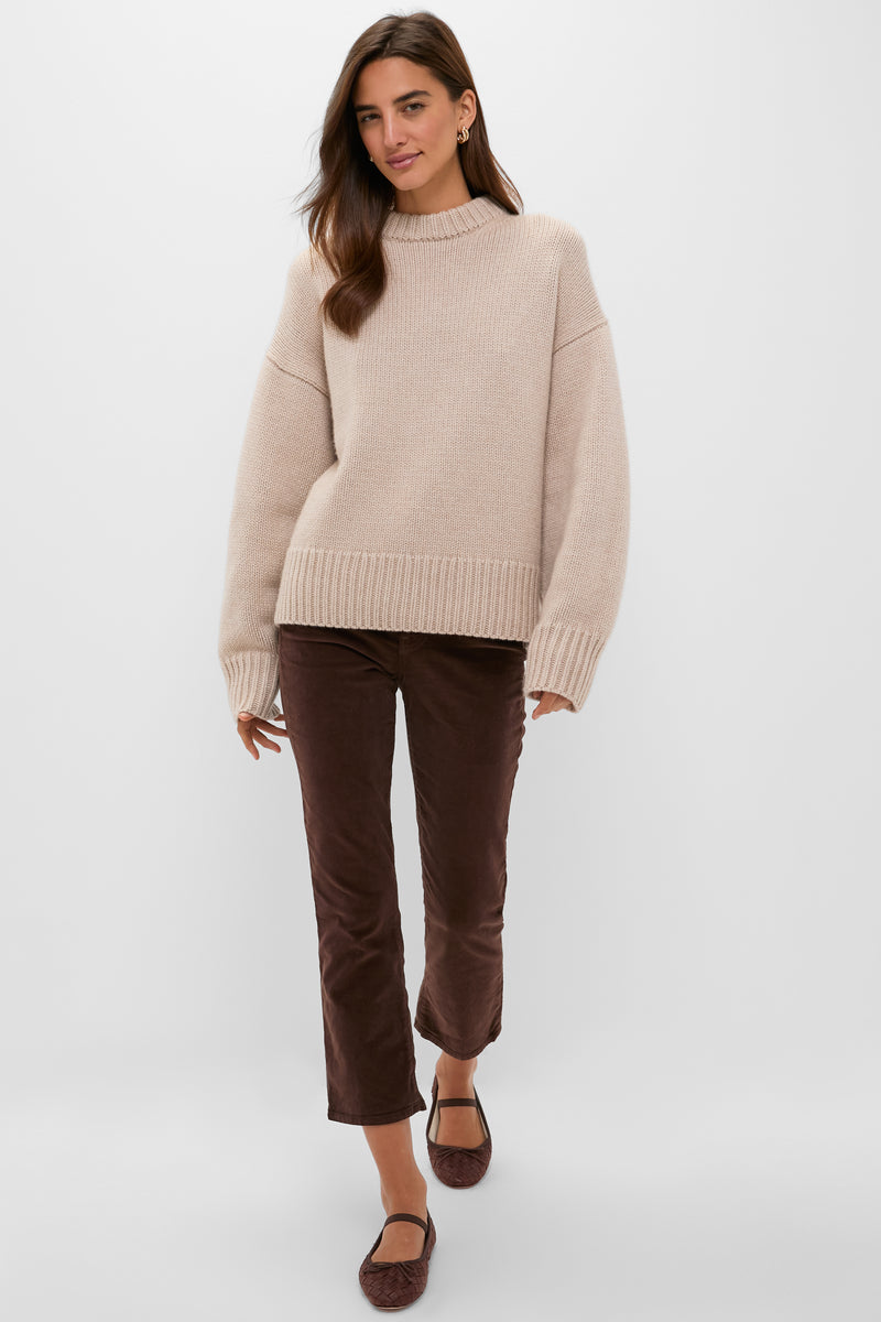 A woman stands against a plain light background wearing a Tuckernuck beige cashmere boxy-fit sweater, brown cropped pants, and brown flats.
