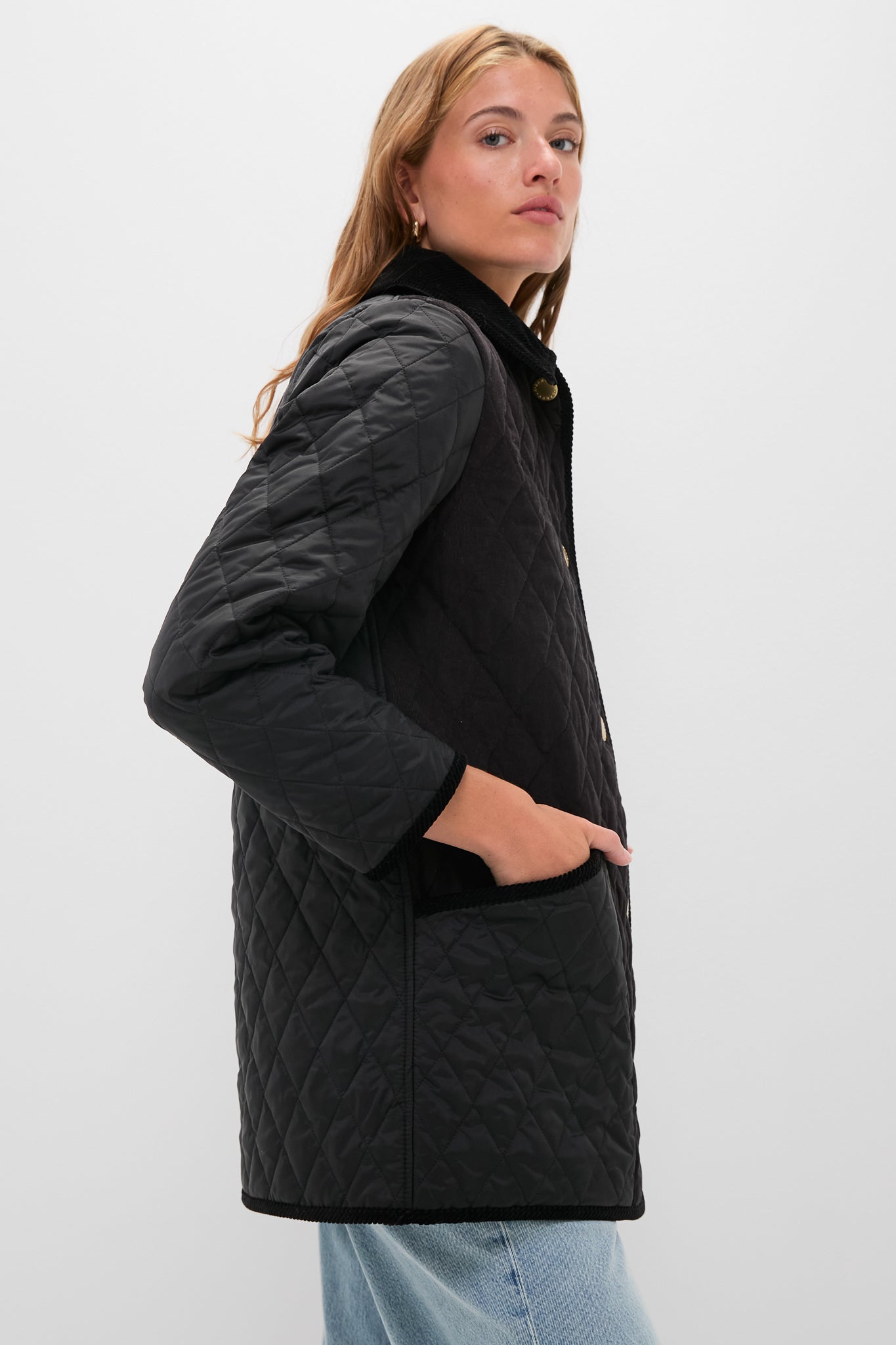 A woman stands against a plain white background wearing a Barbour black jacket with front pockets and light blue jeans.