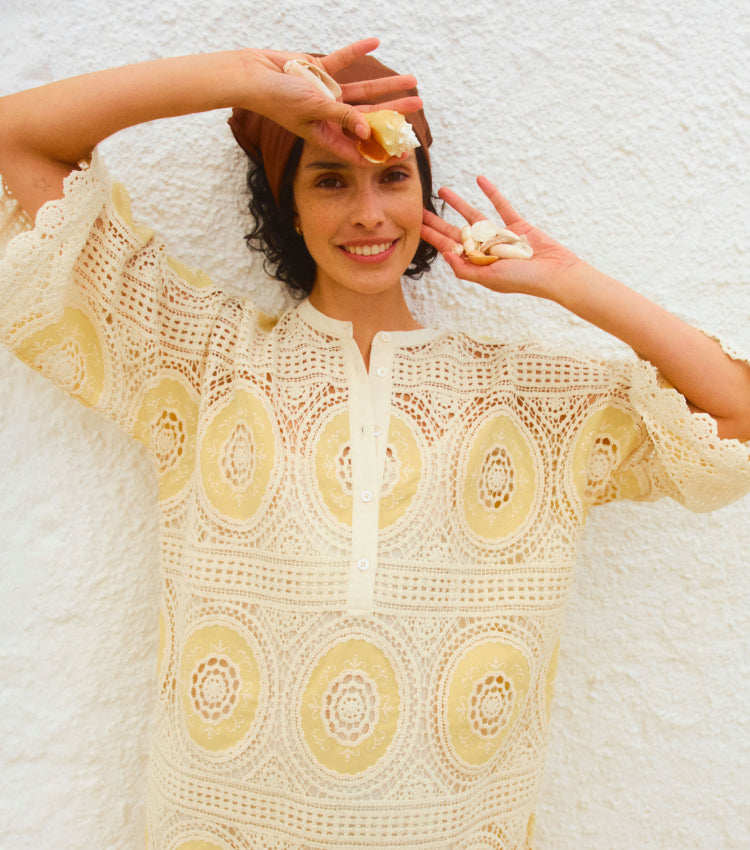 Woman holding three shells in front of her face in a crochet mini dress with yellow accents. 