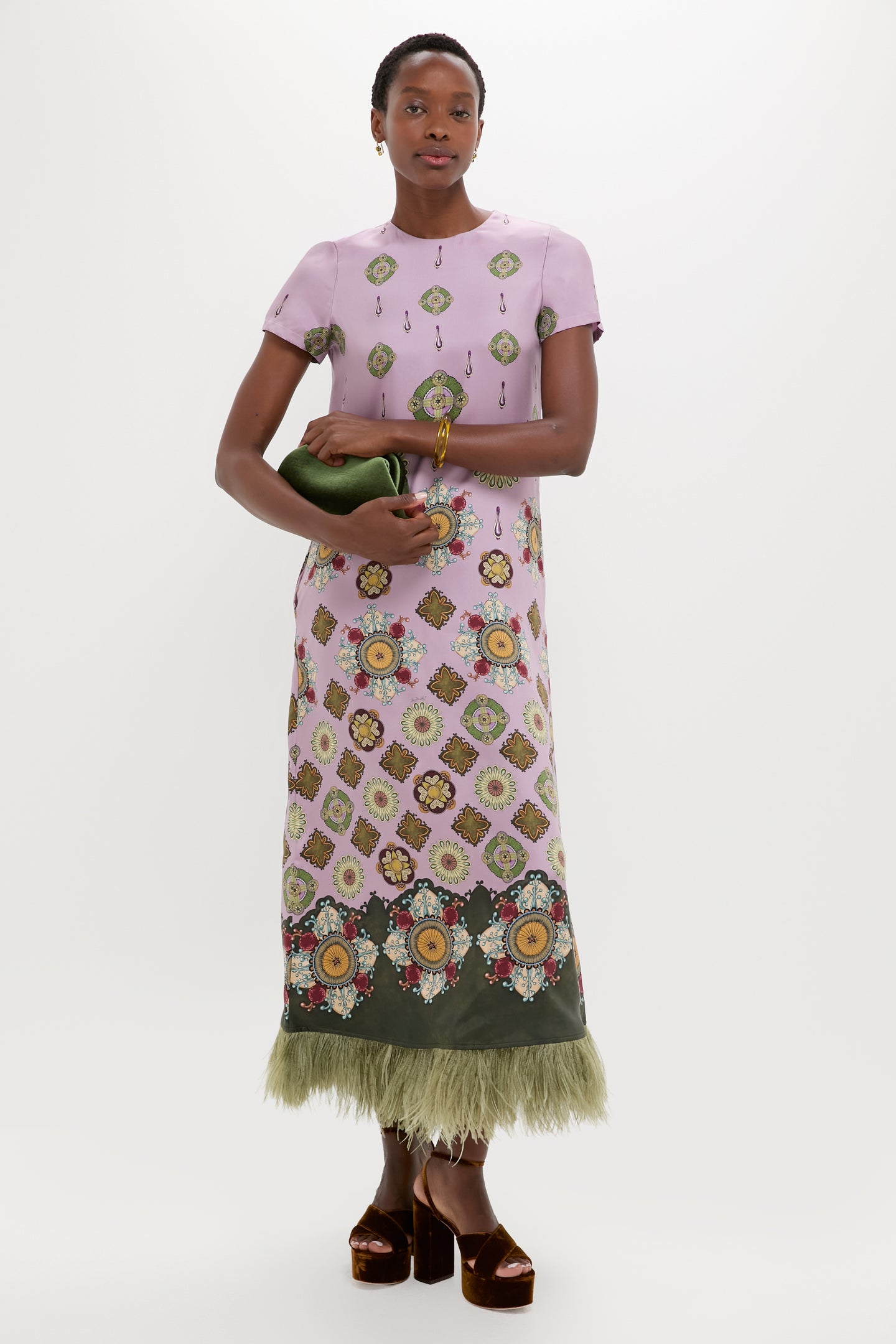 A person wears a La DoubleJ pink silk twill short-sleeve dress with a patterned design and green feather trim, accessorized with a green clutch and brown platform shoes, standing against a white background—ideal for events.