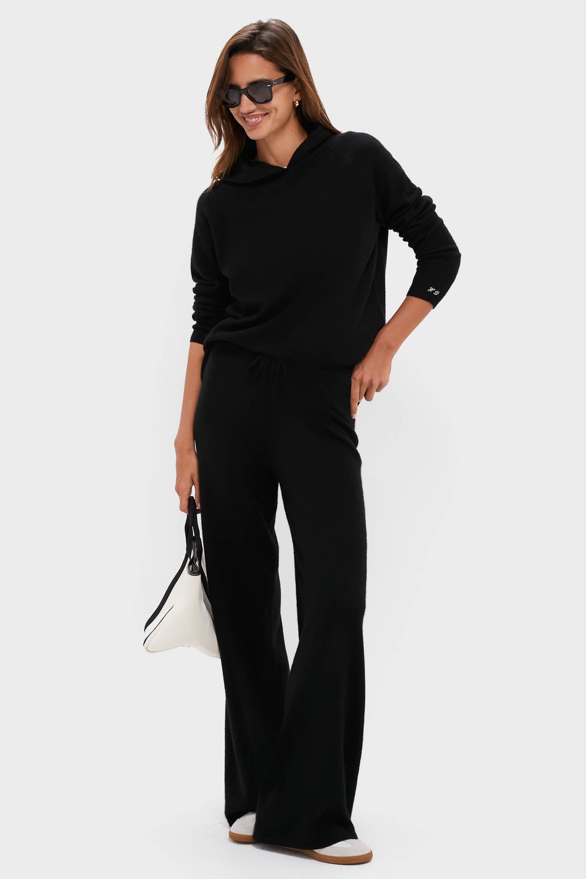 A woman wears a matching black cashmere set with long sleeves and wide-leg pants, styled with sunglasses, a white bag, and light-colored slip-on shoes.
