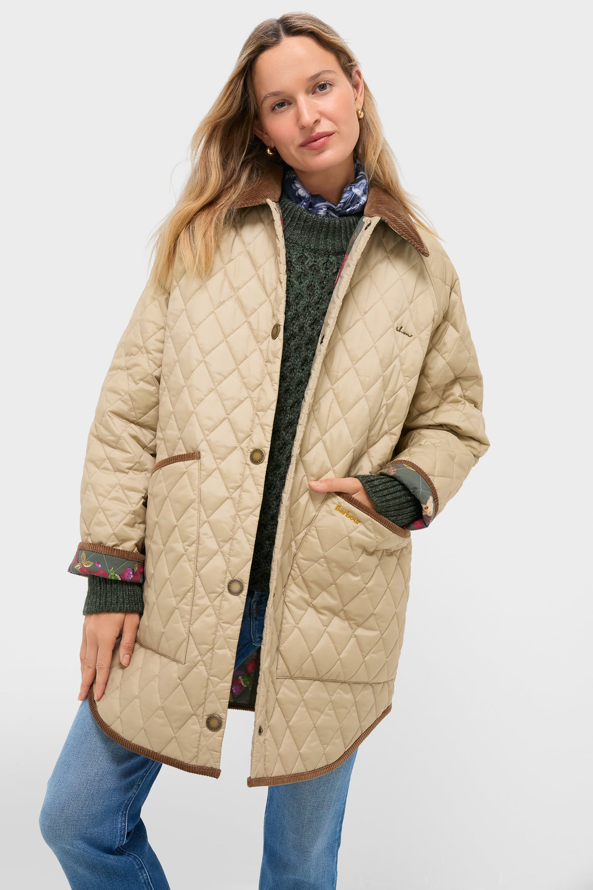 A woman wears the Barbour x Tuckernuck Miley quilt jacket, a beige diamond quilt style with brown trim, over a green sweater and blue jeans, standing with one hand in her pocket against a plain background.