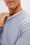 A person wearing a super-soft, eco-friendly light blue textured Marine Layer sweater holds the collar with one hand; only their lower face, neck, and upper torso are visible.