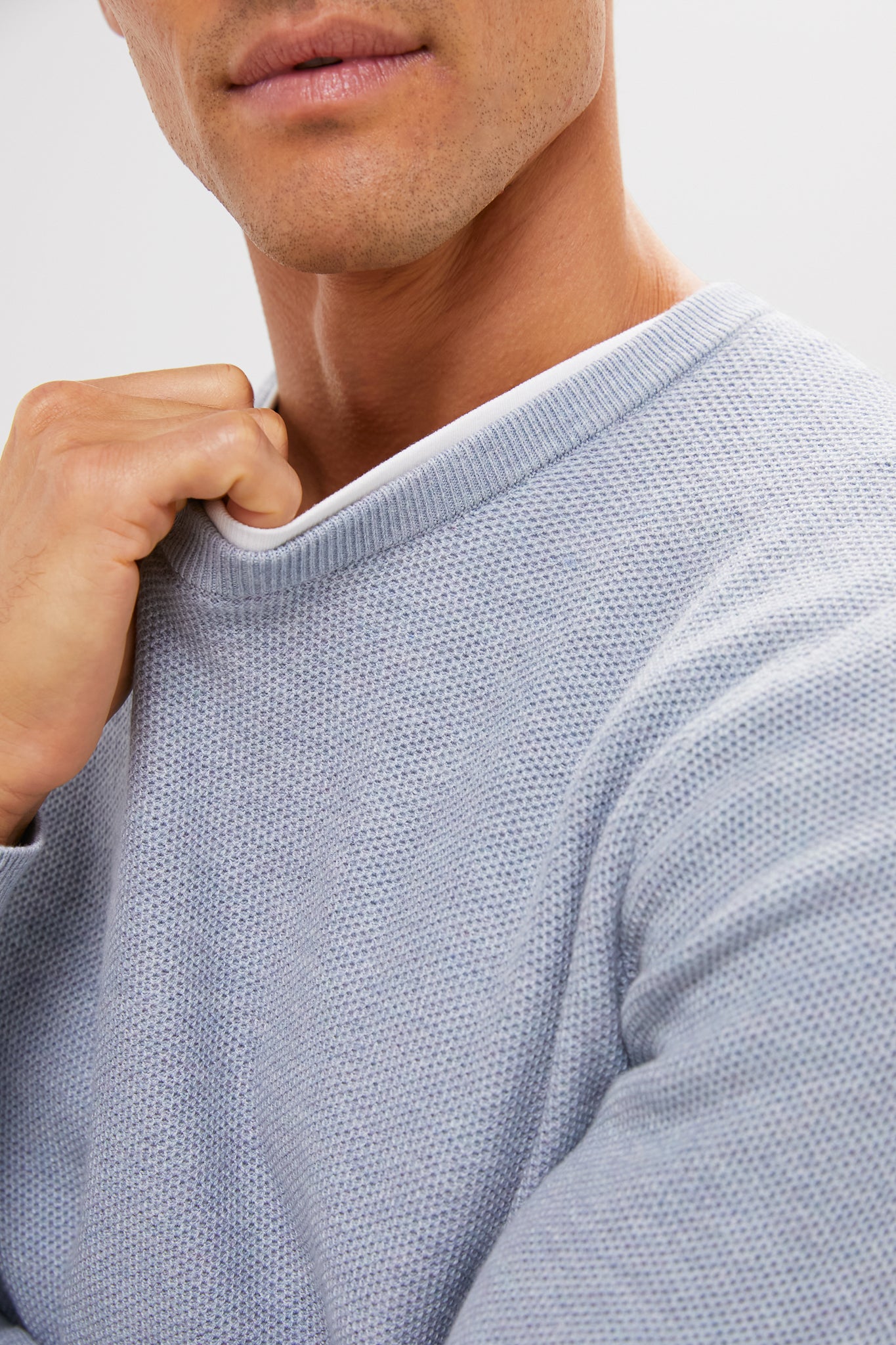 A person wearing a super-soft, eco-friendly light blue textured Marine Layer sweater holds the collar with one hand; only their lower face, neck, and upper torso are visible.