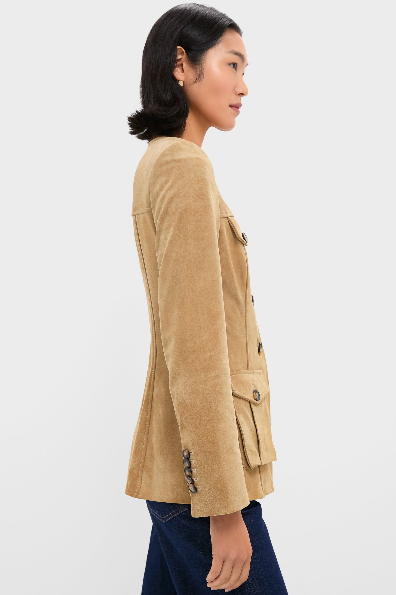 A woman stands in profile against a white background wearing a slim-fit tan suede blazer by Veronica Beard, featuring button details and flap pockets.