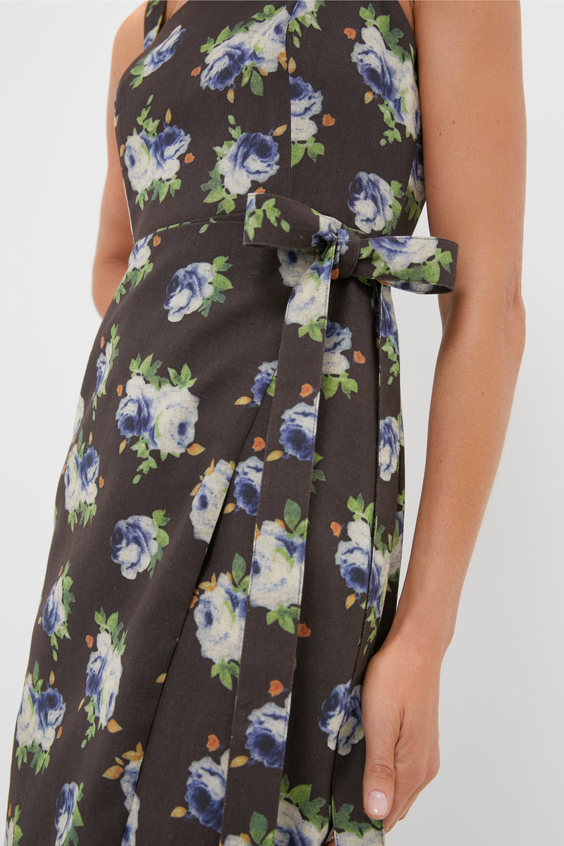 A close-up of a Hyacinth House blue floral midi dress with a waist tie, shown against a dark background; part of a womans arm and hand is visible, emphasizing the elegant wrap style.
