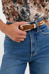 A person wearing Petit Moments blue jeans, a horse-print shirt, and a chocolate leather western belt with goldtone hardware, one hand resting on the belt.