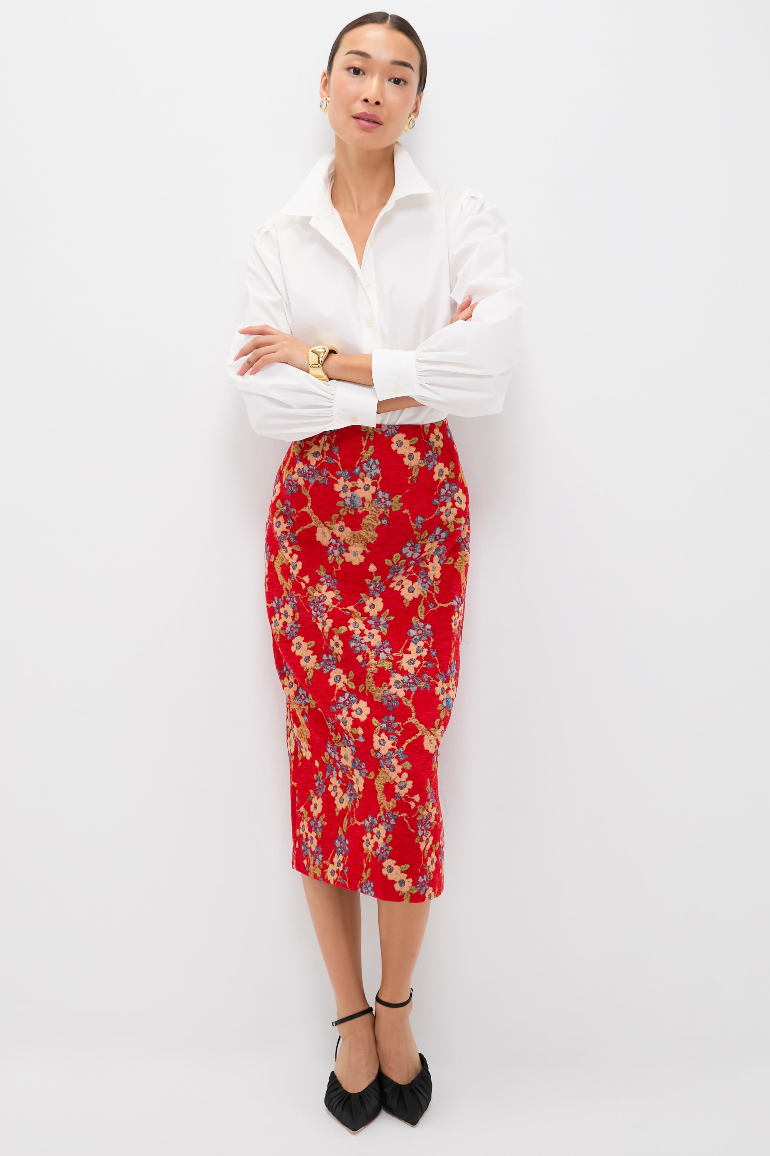 A person poses against a white background wearing a Tuckernuck white collared shirt, a chic floral jacquard skirt, black pointed shoes, and a gold watch, arms crossed.