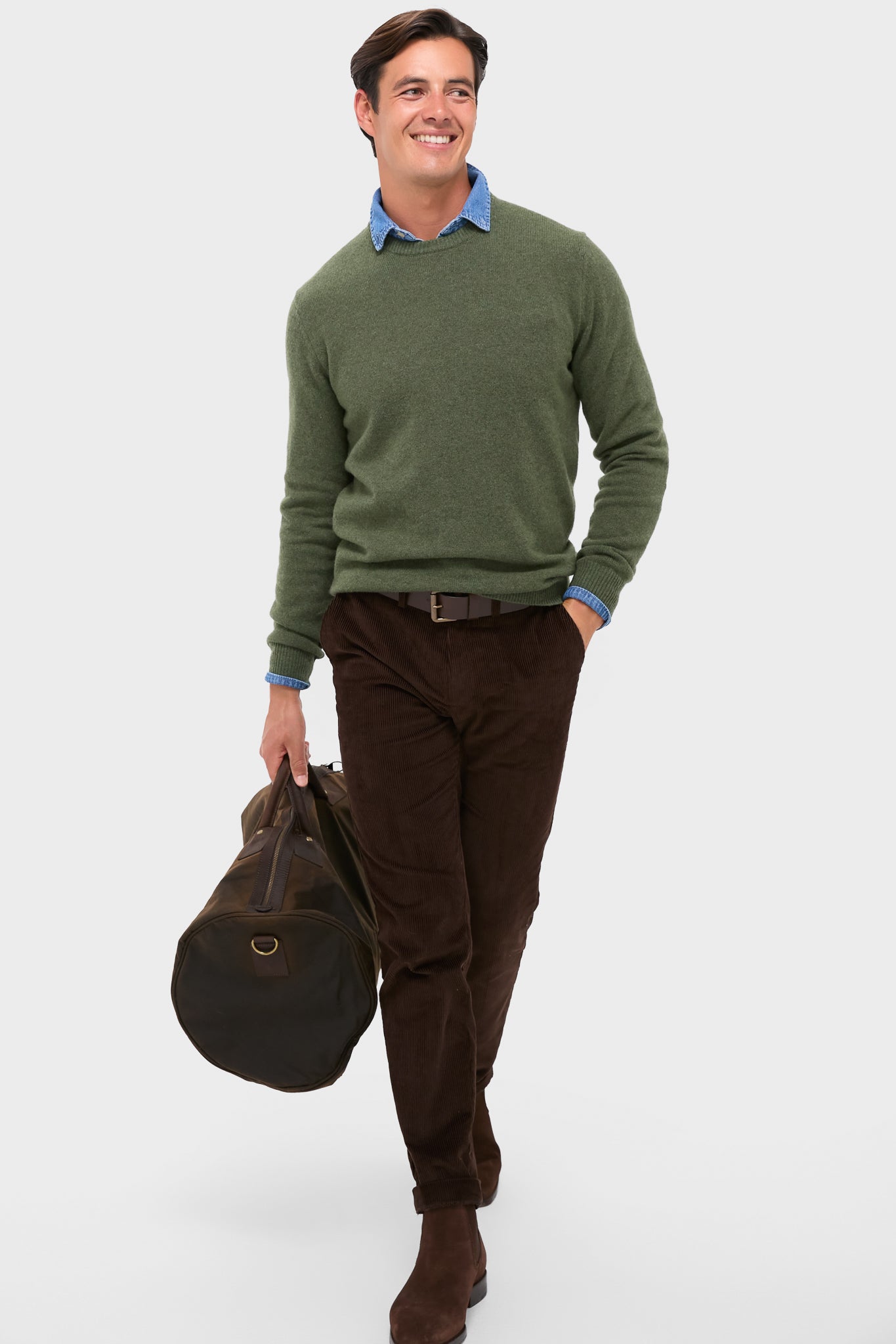 Model wearing Olive Essential Patch Crew Neck Sweater, layered over a chambray shirt, paired with corduroy trousers. Carries a rugged duffel, exuding classic, effortless style.