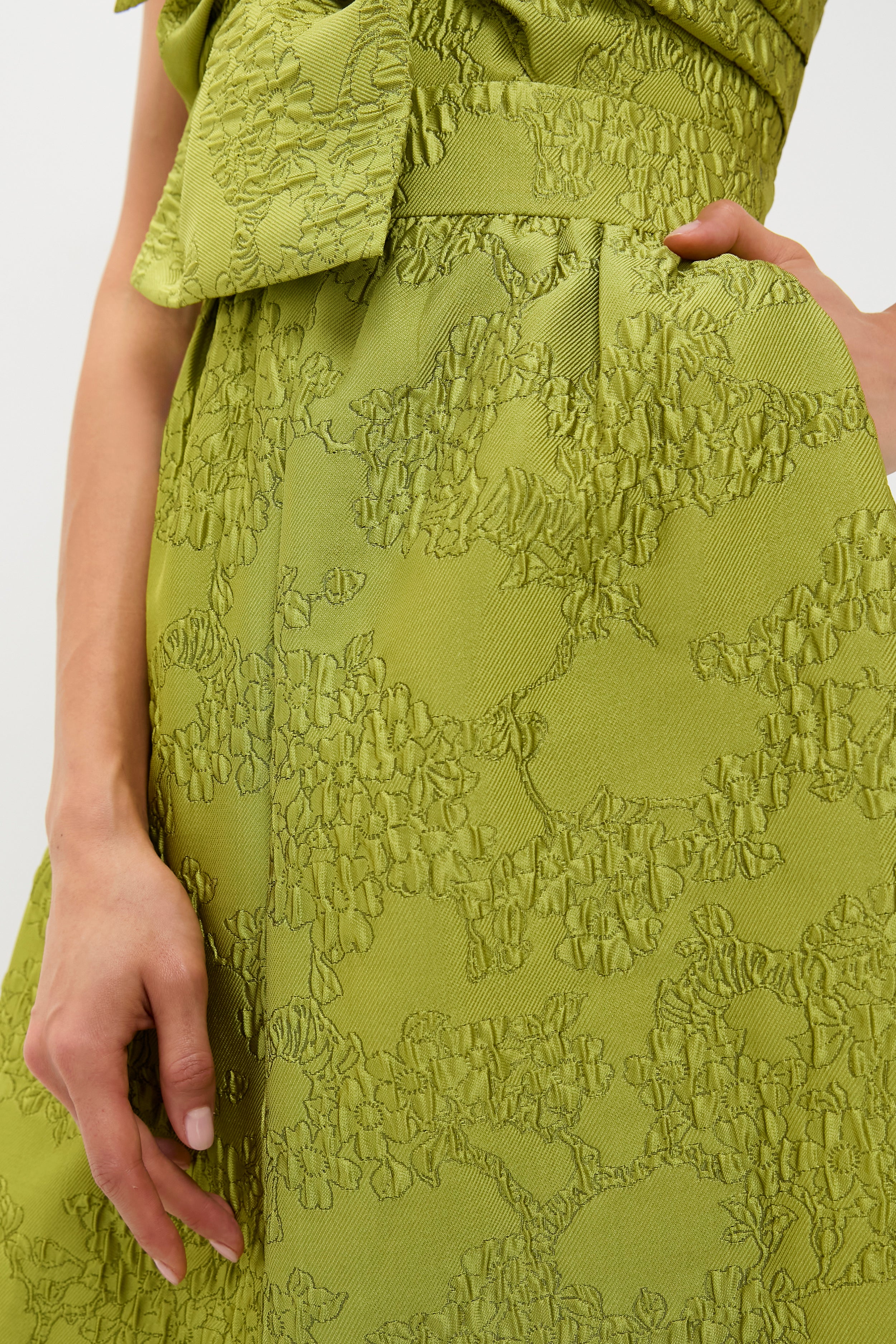 Close-up of a person wearing a textured, light green jacquard gown with an embossed floral pattern by Tuckernuck, showing part of the upper body and a hand resting on the hip.