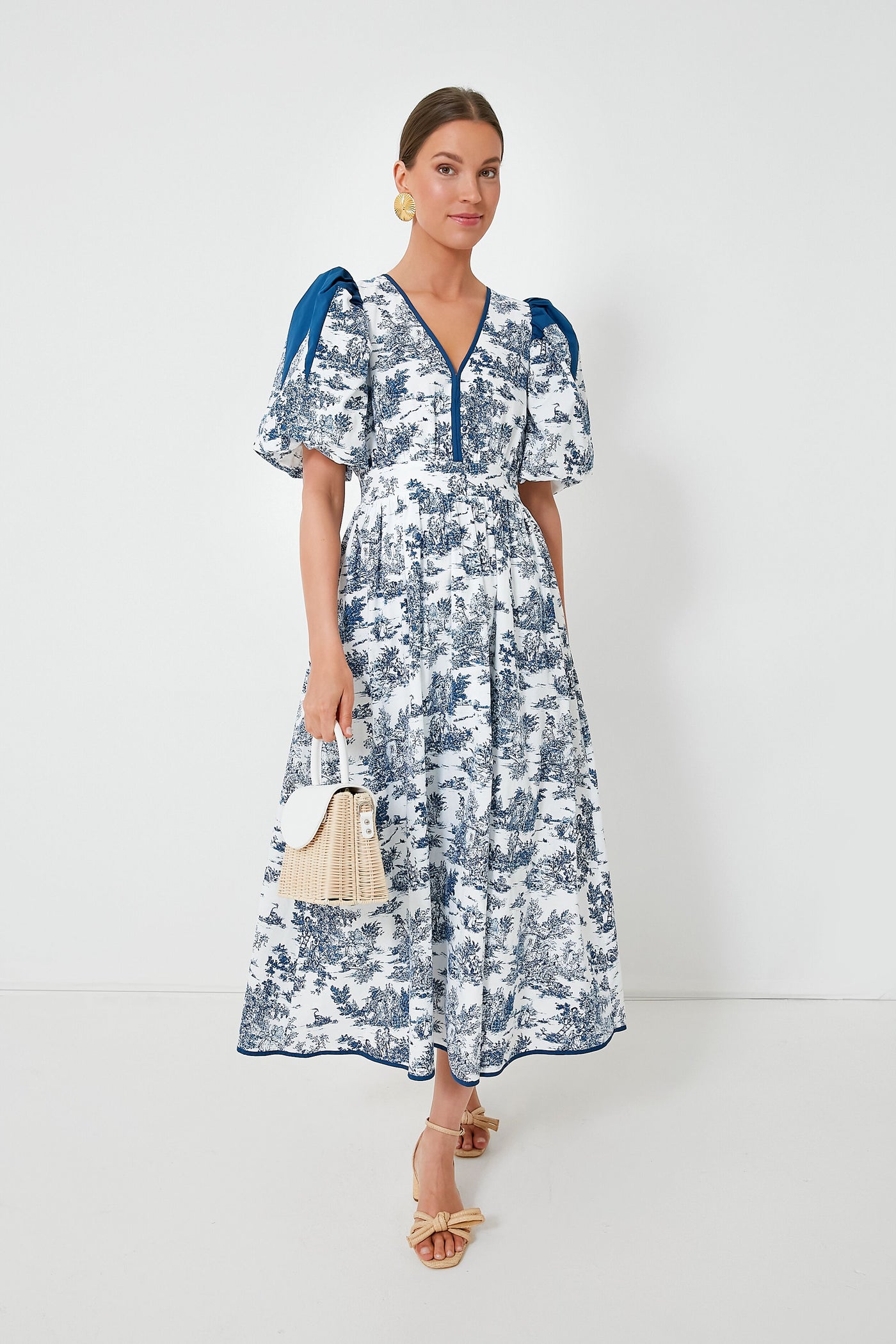 A woman wears a blue and white Hyacinth House Reynolds Dress with puffed sleeves, carrying a small handbag and wearing bow sandals.