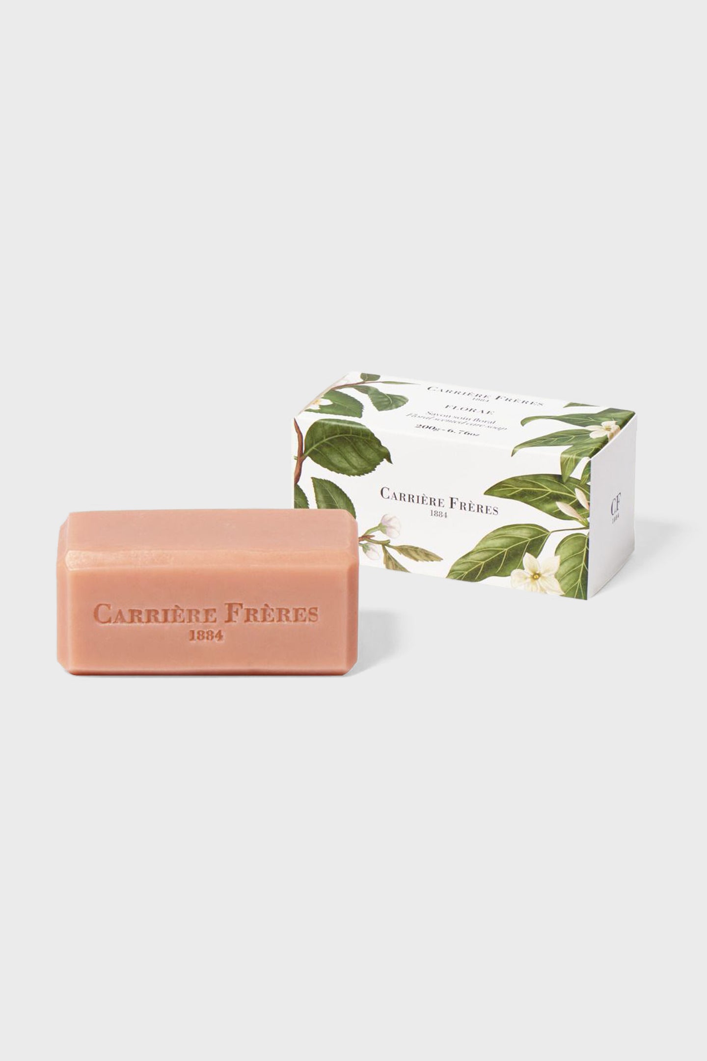 A pink rectangular vegan soap bar embossed with Carrière Frères 1884 sits beside a floral-printed white Carrière Frères box, emphasizing natural ingredients for gentle cleansing.
