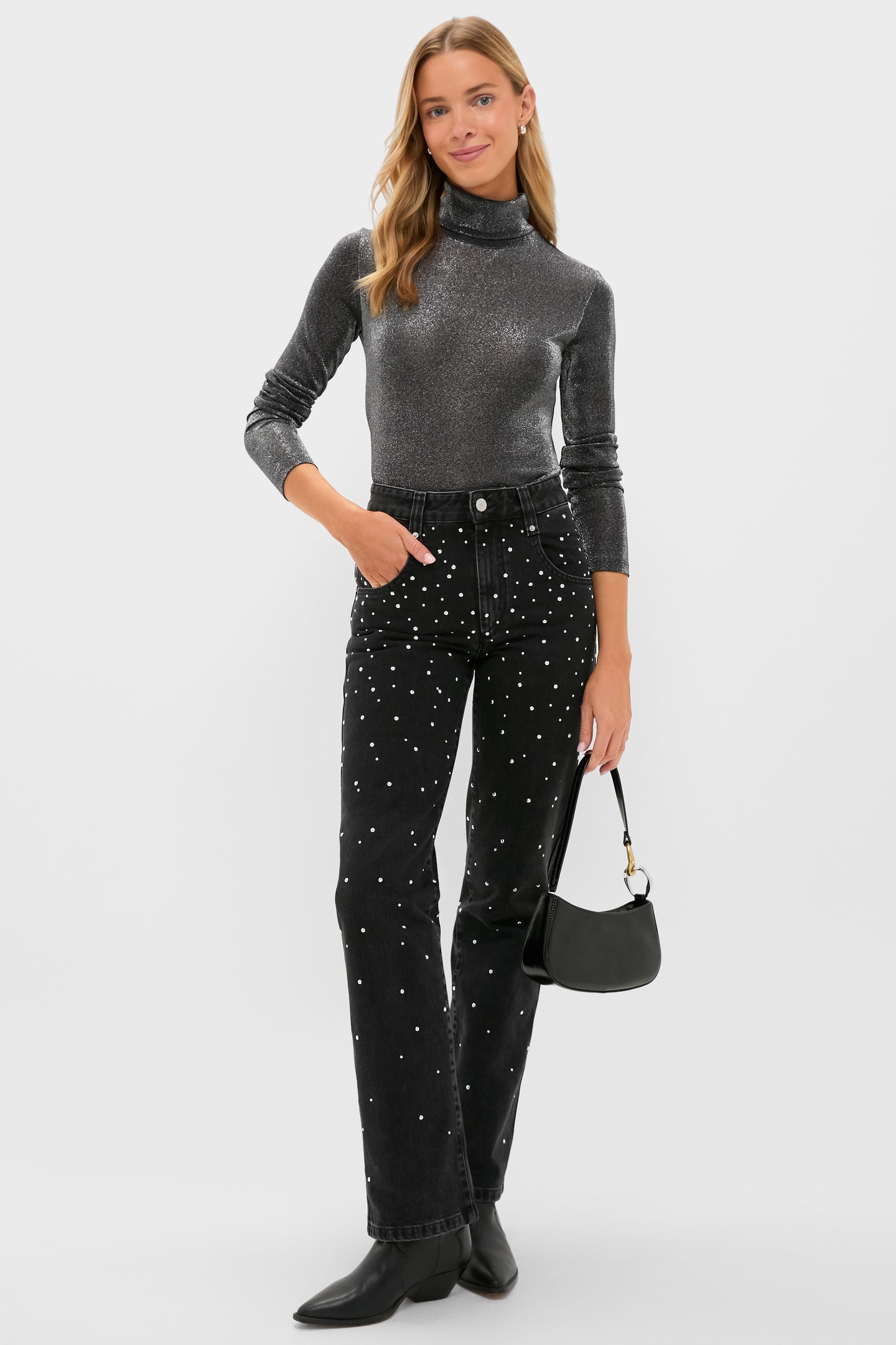 A woman stands smiling in an Isabel Marant Etoile metallic gray jersey top with gathered details, black polka dot jeans, black boots, and a small black handbag.