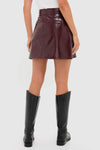 Back view of a person in the Oxblood Meeghan Mini Skirt, a glossy, high-waisted A-line with a sleek back zipper, paired with knee-high black boots.