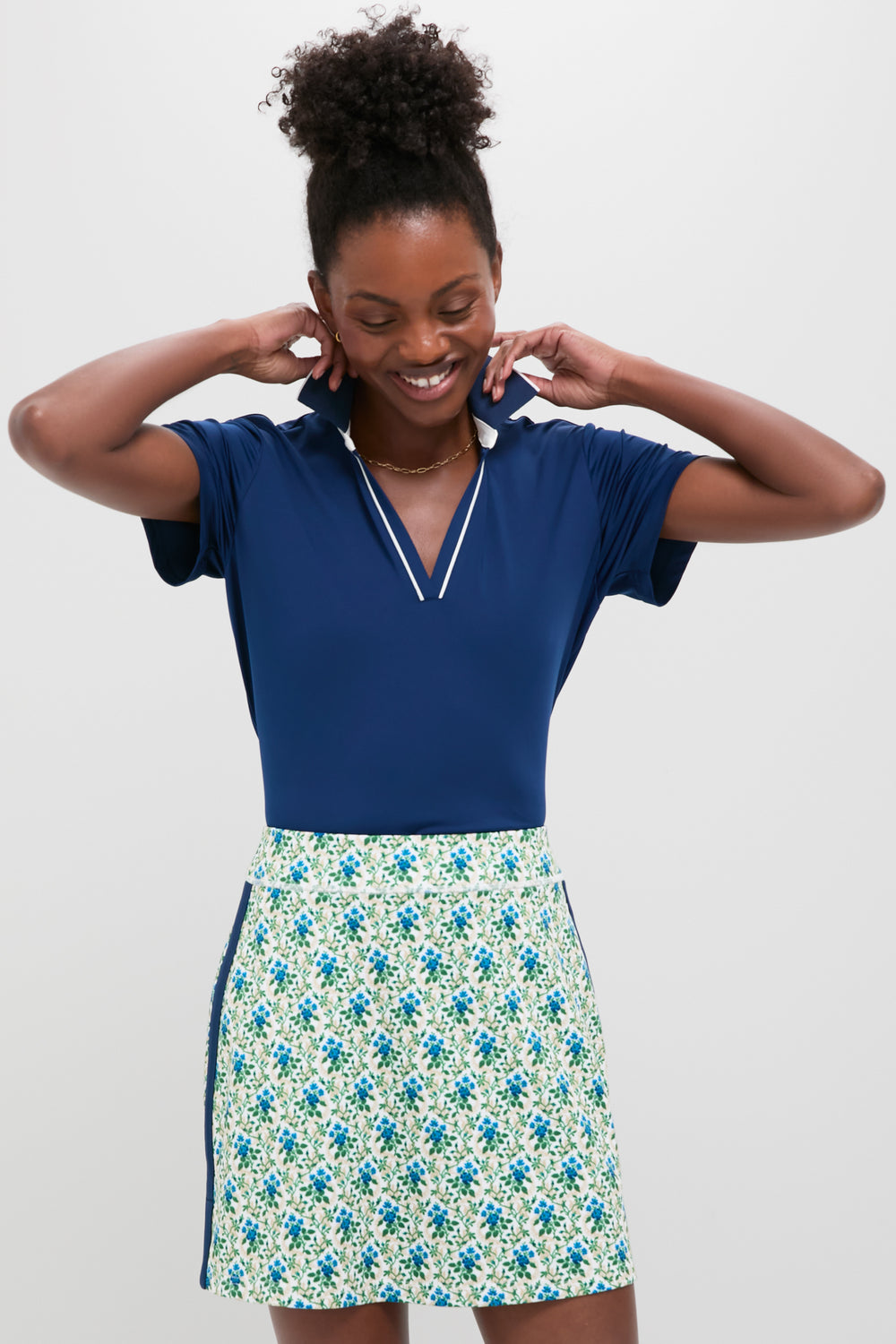 A woman smiles, wearing a Tnuck Sport navy Short Sleeve Polo with SPF Protection and a white skirt featuring a blue and green floral print.